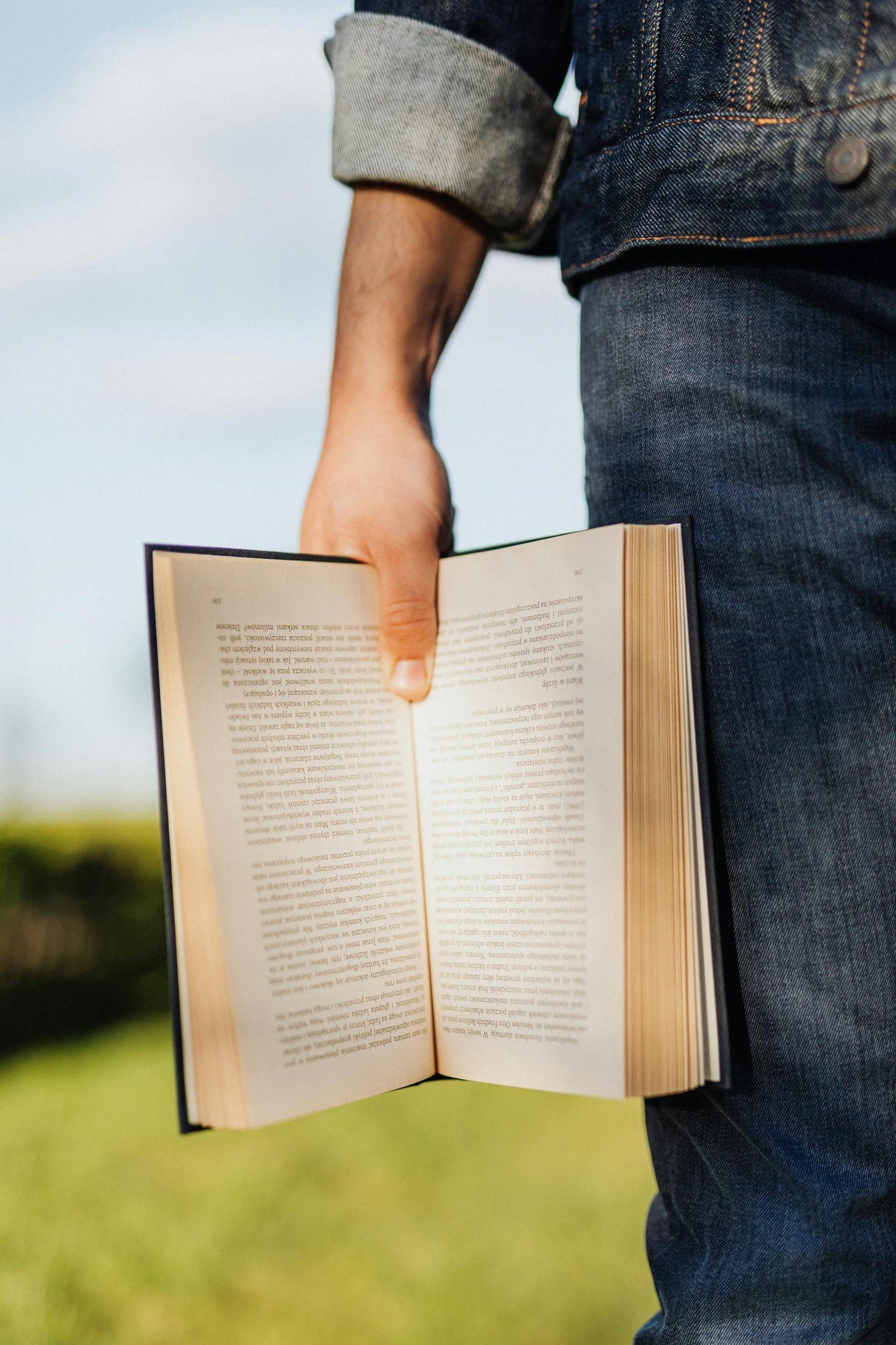 Crop unrecognizable male student in casual outfit standing with book in hand on green spring meadow with sunshine lighting up pages of novel