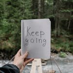 Person Holding a Notebook with a Text "Keep Going" on the Background of a River in a Forest