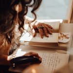 Crop anonymous female with dark hair sitting near window at retro writing cabinet while taking notes with feather in vintage store