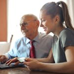 Young woman in casual clothes helping senior man in formal shirt with paying credit card in Internet using laptop while sitting at table